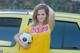 A young woman holding a soccer ball in front of a yellow car.