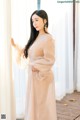 a woman standing in front of a window wearing a beige dress