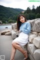 A woman in a blue sweater and white dress sitting on a stone wall.